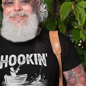 K&ouml;nnte beinhalten: Schwarzes T-Shirt mit einer wei&szlig;en Grafik einer Person, die von einem Boot aus angelt, mit dem Text "HOOKIN' AINT' EASY".