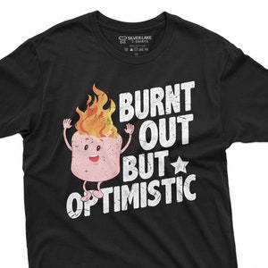 Puede incluir: Camiseta negra con un gráfico blanco de un malvavisco quemado con una cara sonriente y el texto "Burnt Out But Optimistic".