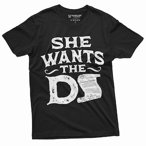 Puede incluir: Camiseta negra con la frase "SHE WANTS THE D" en blanco desgastado. Debajo, un pergamino con "Declaration of Independence" y la fecha "1776". La camiseta es de Silver Lake T-Shirts.