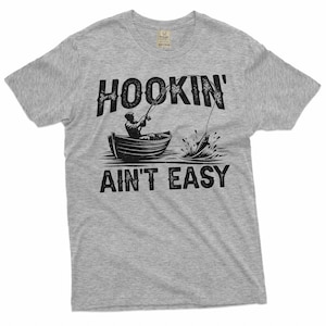 K&ouml;nnte beinhalten: Graues T-Shirt mit einer schwarzen Grafik eines Fischers in einem Boot mit dem Text "HOOKIN' AINT' EASY".