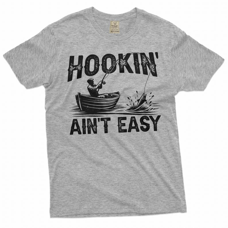 K&ouml;nnte beinhalten: Graues T-Shirt mit einer schwarzen Grafik eines Fischers in einem Boot mit dem Text "HOOKIN' AINT' EASY".