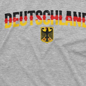 May include: A gray t-shirt with the word "DEUTSCHLAND" printed in black, red, and yellow letters. A black eagle with red claws and beak is centered below the word, surrounded by a yellow circle with a red border.