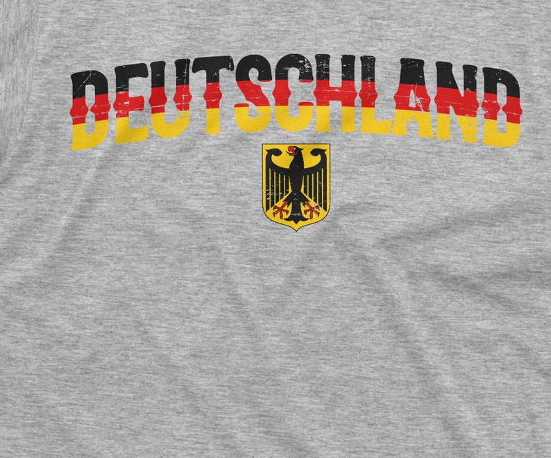 May include: A gray t-shirt with the word "DEUTSCHLAND" printed in black, red, and yellow letters. A black eagle with red claws and beak is centered below the word, surrounded by a yellow circle with a red border.