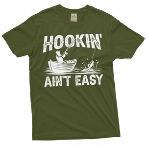 K&ouml;nnte beinhalten: Ein gr&uuml;nes T-Shirt mit einer wei&szlig;en Grafik einer Person, die von einem Boot aus angelt. Der Text auf dem Shirt lautet "Hookin' Ain't Easy".