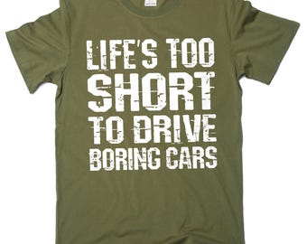 May include: Olive green t-shirt with the white text "LIFE'S TOO SHORT TO DRIVE BORING CARS". The shirt has a crew neck and short sleeves. The text is in a bold, distressed font.