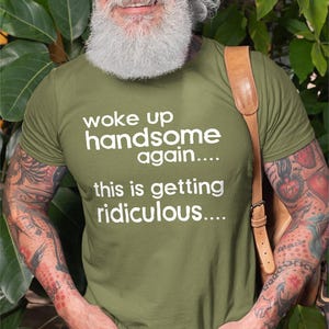 Könnte beinhalten: Ein olivgrünes Kurzarm-T-Shirt mit dem weißen Text "woke up handsome again.... this is getting ridiculous...." auf der Brust. Das T-Shirt wird mit einer blauen Jeans und einem beigen Schulterriemen aus Leder getragen.