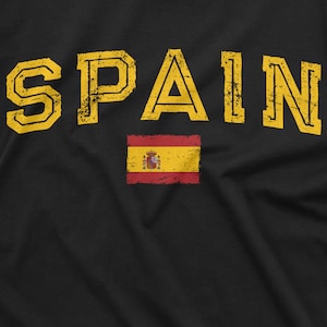 K&ouml;nnte beinhalten: Ein schwarzes T-Shirt mit dem Wort "SPAIN" in gelber, abgenutzter Schrift. Unter dem Wort befindet sich eine kleine rot-gelbe spanische Flagge.