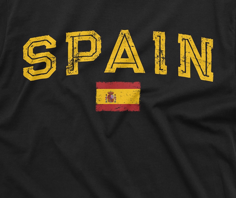 K&ouml;nnte beinhalten: Ein schwarzes T-Shirt mit dem Wort "SPAIN" in gelber, abgenutzter Schrift. Unter dem Wort befindet sich eine kleine rot-gelbe spanische Flagge.