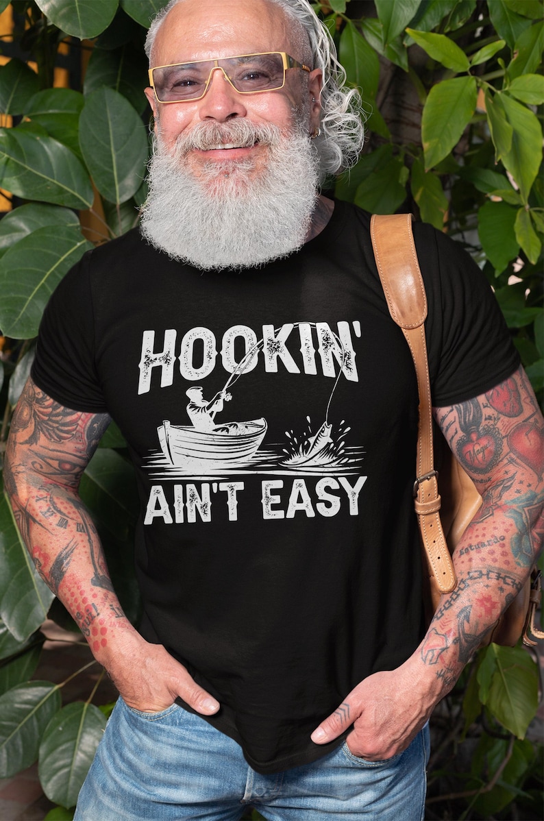 K&ouml;nnte beinhalten: Schwarzes T-Shirt mit einer wei&szlig;en Grafik einer Person, die von einem Boot aus angelt, mit dem Text "HOOKIN' AINT' EASY".