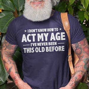 Op de afbeelding: Een marineblauw t-shirt met witte tekst die luidt "★ I DON'T KNOW HOW TO ★ ACT MY AGE - I'VE NEVER BEEN - THIS OLD BEFORE"