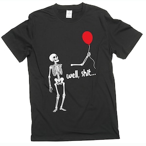 Può includere: Una t-shirt nera a maniche corte con una grafica bianca di uno scheletro che guarda verso un braccio scheletrico staccato che tiene un palloncino rosso. Il testo "well, shit..." è stampato in lettere bianche sotto il braccio, al centro.
