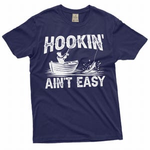 K&ouml;nnte beinhalten: Ein marineblaues T-Shirt mit einer wei&szlig;en Grafik einer Person, die von einem Boot aus angelt. Der Text "HOOKIN' AINT EASY" ist in Wei&szlig; auf das Shirt gedruckt.