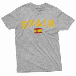 K&ouml;nnte beinhalten: Ein graues T-Shirt mit dem Wort "SPAIN" in gelber Farbe und einem abgenutzten Look. Die spanische Flagge ist unter dem Wort gedruckt.