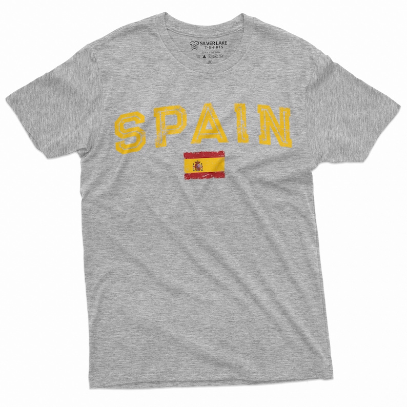 K&ouml;nnte beinhalten: Ein graues T-Shirt mit dem Wort "SPAIN" in gelber Farbe und einem abgenutzten Look. Die spanische Flagge ist unter dem Wort gedruckt.