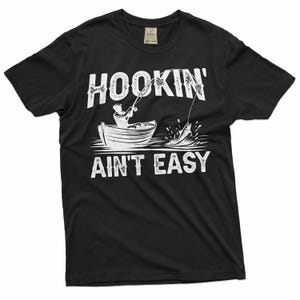 K&ouml;nnte beinhalten: Schwarzes T-Shirt mit einer wei&szlig;en Grafik einer Person, die von einem Boot aus angelt, mit dem Text "HOOKIN' AINT' EASY".