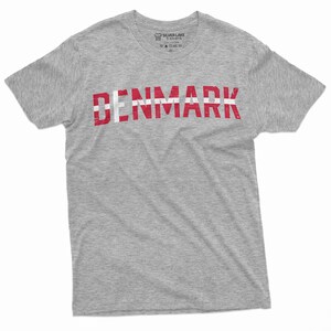 May include: A gray t-shirt with the word "DENMARK" printed in red and white letters. The letters are stylized to look like the Danish flag.