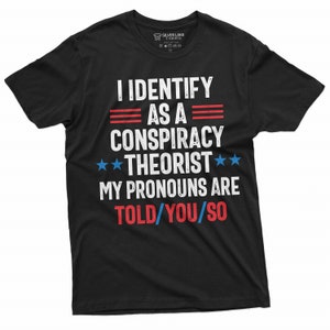 May include: Black t-shirt with a white graphic that says "I identify as a conspiracy theorist. My pronouns are told/you/so". The graphic also includes three red stripes, two blue stars, and the text is in a distressed font.