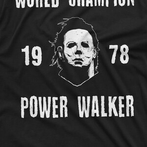 May include: Black and white graphic t-shirt featuring a white image of a masked figure and the text "WORLD CHAMPION 19 78 POWER WALKER".