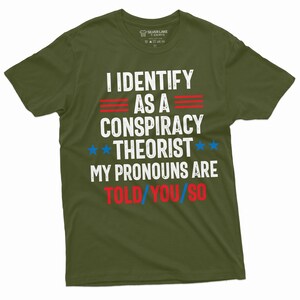 May include: A green t-shirt with the text "I identify as a conspiracy theorist. My pronouns are told/you/so" in white, red, and blue lettering. The text is in a distressed font style.