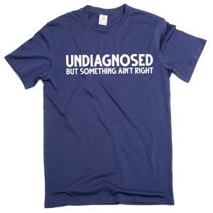 May include: Navy blue t-shirt with the white text "UNDIAGNOSED BUT SOMETHING AIN'T RIGHT". The shirt is a crew neck style and appears to be made of a soft, comfortable material. The text is centered on the front of the shirt.