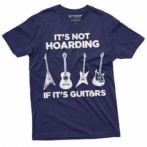 May include: Navy blue t-shirt with white text that reads "It's not hoarding if it's guitars". The shirt features a graphic of five guitars, including an acoustic guitar, an electric guitar, and a bass guitar.