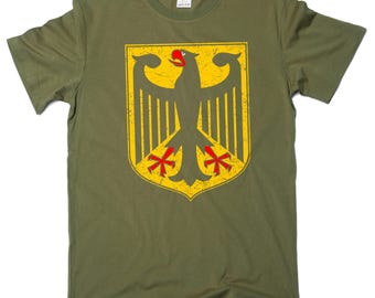 May include: Olive green t-shirt featuring a distressed yellow and black German eagle emblem. The emblem is a shield shape with a central eagle design and red accents. The shirt is made of a soft material.