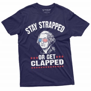 Peut inclure: Un t-shirt bleu marine avec un graphique blanc de George Washington portant des lunettes de soleil avec un motif de drapeau américain. Le texte "Stay Strapped or Get Clapped" est imprimé sur le t-shirt.