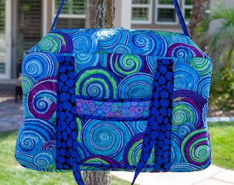 May include: A vibrant, quilted tote bag with a swirling, abstract pattern in shades of blue, green, and purple. The bag has a solid blue strap and accents, and is hanging from a tree branch. The bag is approximately 16 inches wide.