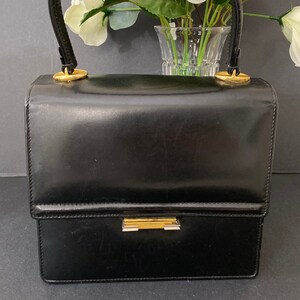 May include: A black leather handbag with a curved handle and gold-toned hardware. The rectangular purse has a clasp closure and a structured design. The bag is set against a backdrop of white and pink flowers in a glass vase.