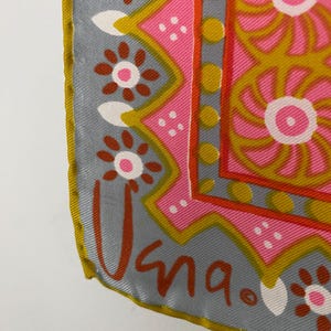 May include: A square silk scarf featuring a vibrant pink center with gold spiral patterns, bordered by a gray band with brown and white floral motifs. The scarf has a gold-colored rolled hem and the signature "Vera" printed in brown in the corner.