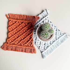 May include: Two handmade macrame coasters, one in orange and one in white, with a small potted cactus in the centre.