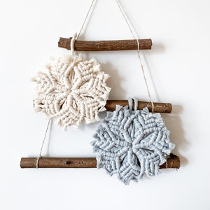 Puede incluir: Una decoración de pared con dos copos de nieve de macramé, uno en crema y otro en azul claro, colgados de listones de madera en una pared blanca.
