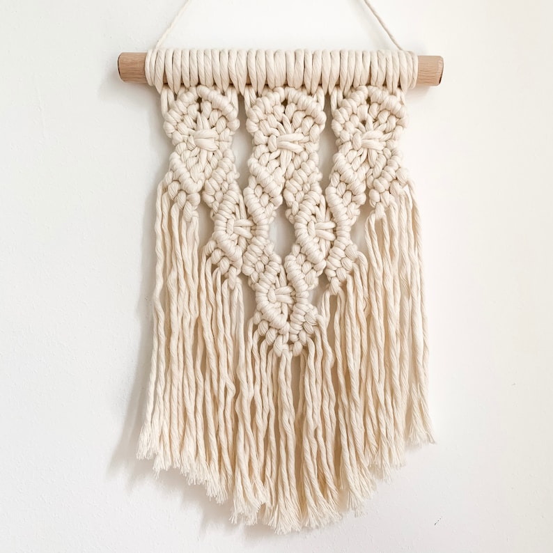 May include: A white macrame wall hanging with a geometric pattern and fringe. The hanging is attached to a wooden dowel.