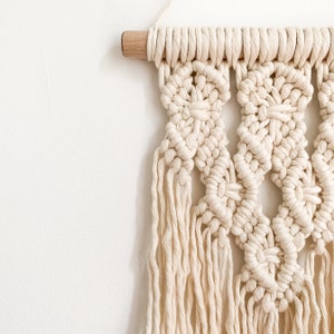 May include: A white macrame wall hanging with a geometric pattern and fringe details. The hanging is attached to a wooden dowel.