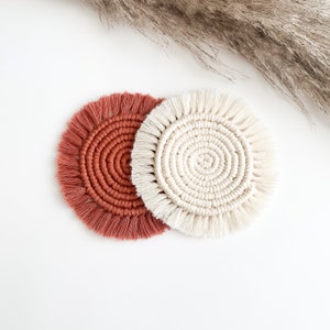 May include: Two round crocheted coasters, one in a terracotta orange color and one in a cream color. Both coasters have a fringe border.