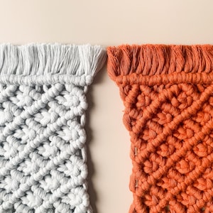 Peut inclure: Deux tentures murales en macram&eacute;, l'une de couleur gris clair et l'autre de couleur orange br&ucirc;l&eacute;. Les deux tentures pr&eacute;sentent un motif en losange et ont une bordure en franges.