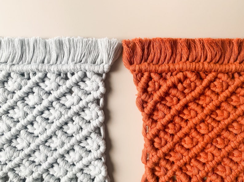 Peut inclure: Deux tentures murales en macram&eacute;, l'une de couleur gris clair et l'autre de couleur orange br&ucirc;l&eacute;. Les deux tentures pr&eacute;sentent un motif en losange et ont une bordure en franges.