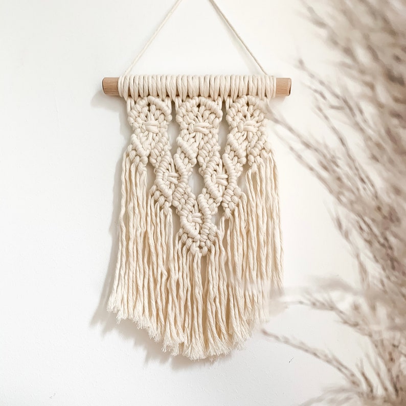 May include: A white macrame wall hanging with a wooden dowel at the top. The hanging features a geometric pattern with fringe details.