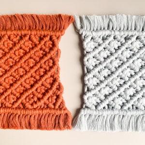 Peut inclure: Deux dessous-de-verre en macram&eacute; faits &agrave; la main, l'un de couleur orange br&ucirc;l&eacute;e et l'autre de couleur gris clair. Les deux dessous-de-verre ont un motif en losange et des franges autour des bords.