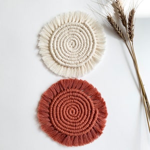 May include: Two handmade macrame coasters, one in white and one in terracotta. The coasters have a circular shape with a fringe border.