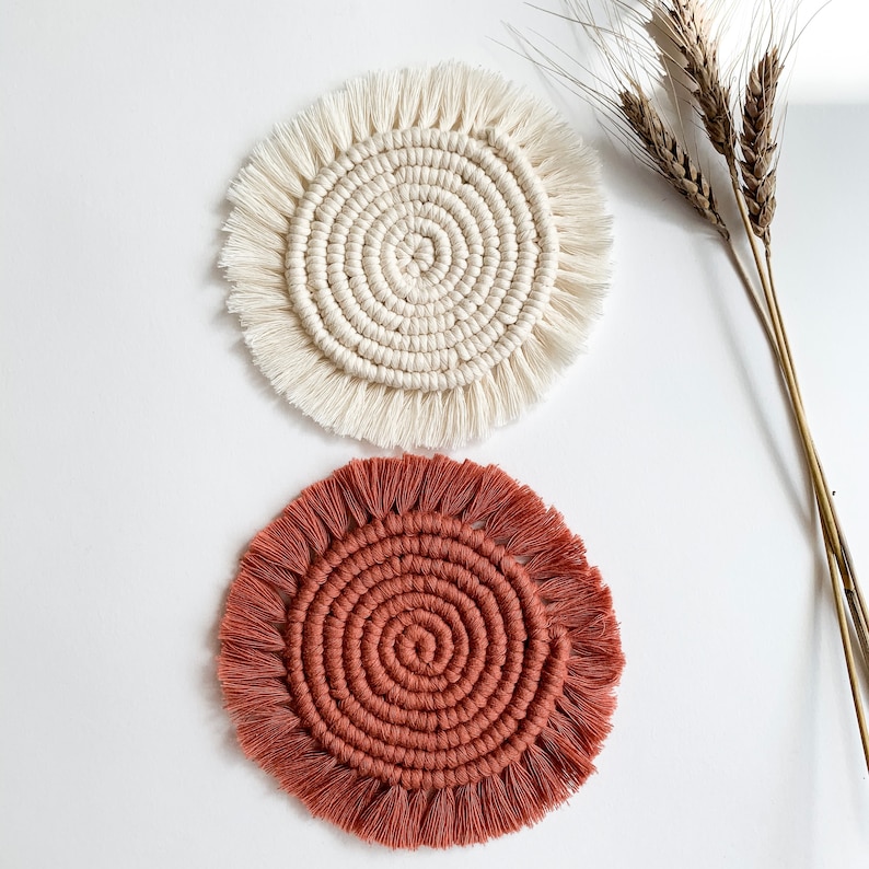 May include: Two handmade macrame coasters, one in white and one in terracotta. The coasters have a circular shape with a fringe border.