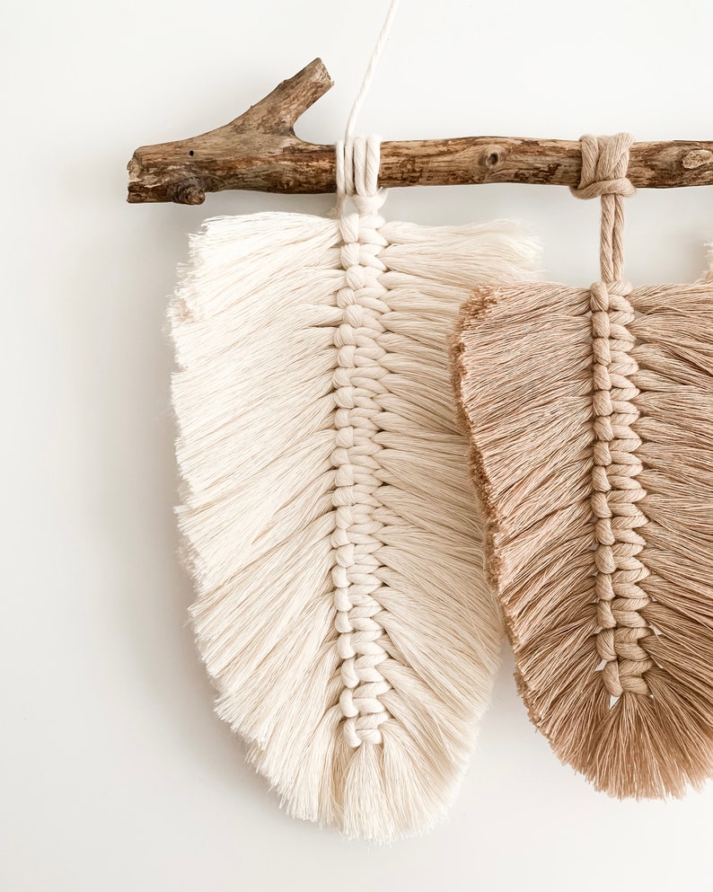 May include: Two macrame feather wall hangings, one white and one brown, are hung from a natural branch. The feathers are made with a knotted rope technique and have a fringe detail.