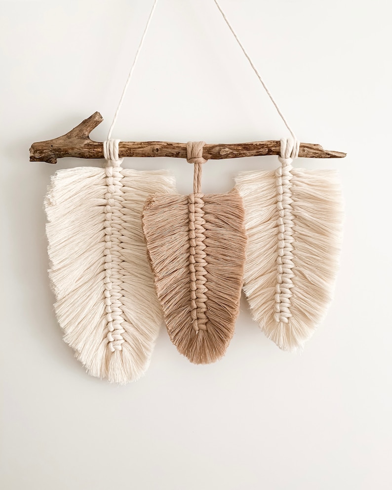 May include: A macrame wall hanging with three feathers, two white and one brown, hanging from a natural branch.