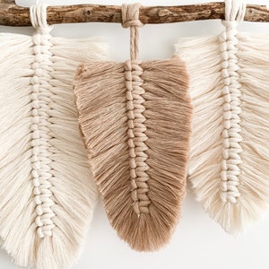 May include: Three macrame feather wall hangings, one brown and two white, are hung on a natural branch. The feathers are made with a knotted rope technique.