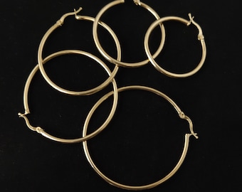 Pode incluir: Quatro brincos de argola dourados de diferentes tamanhos, dispostos em círculo sobre um fundo preto.