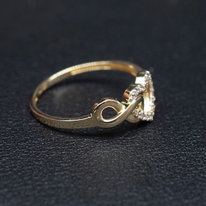 May include: A gold ring with a heart-shaped design featuring a double infinity symbol. The ring is adorned with small, clear stones.