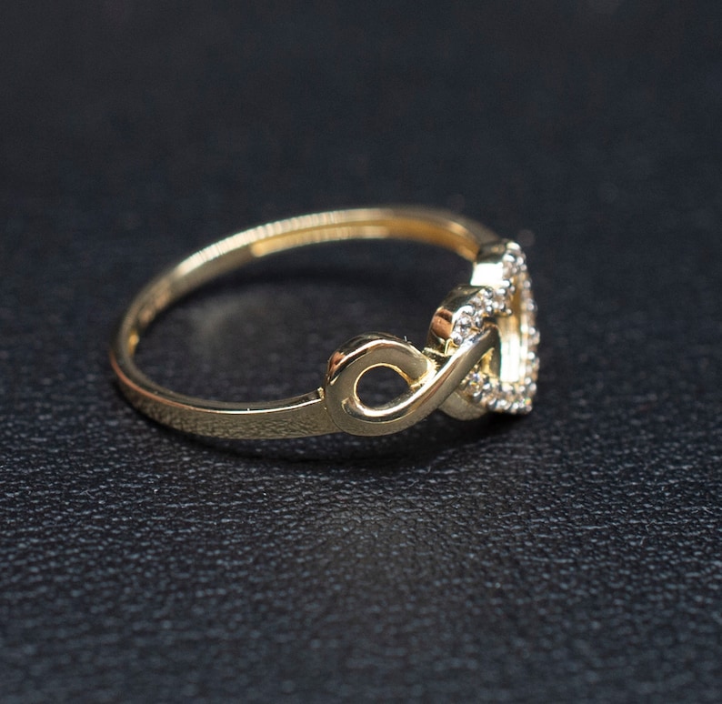 May include: A gold ring with a heart-shaped design featuring a double infinity symbol. The ring is adorned with small, clear stones.