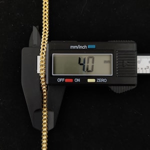 May include: A gold chain is measured with a digital caliper. The caliper reads 4.0 mm.