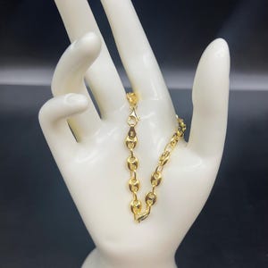 K&ouml;nnte beinhalten: Ein goldenes Kettenarmband mit einem Ankerketten-Design. Das Armband befindet sich an einer wei&szlig;en Mannequin-Hand.