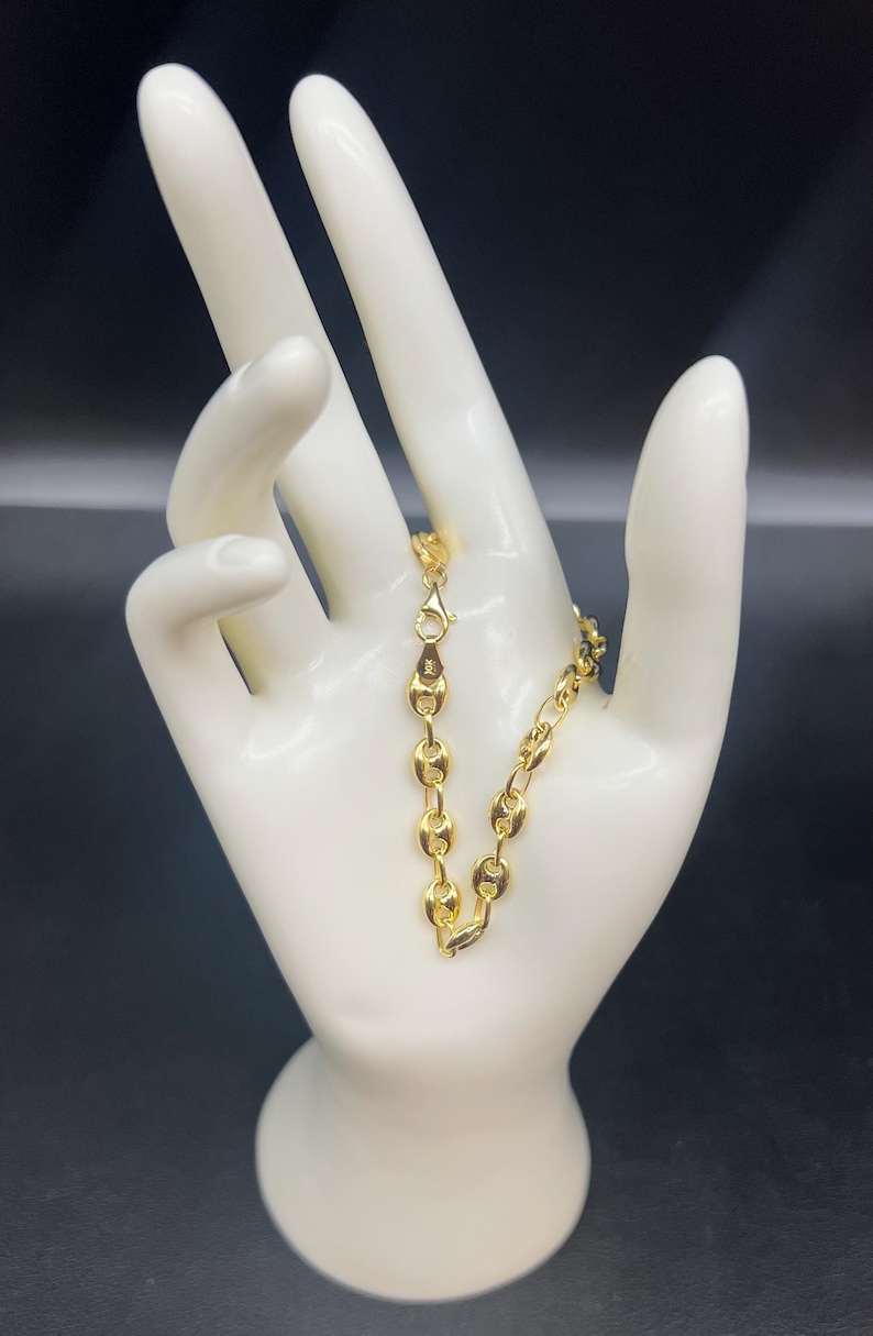K&ouml;nnte beinhalten: Ein goldenes Kettenarmband mit einem Ankerketten-Design. Das Armband befindet sich an einer wei&szlig;en Mannequin-Hand.